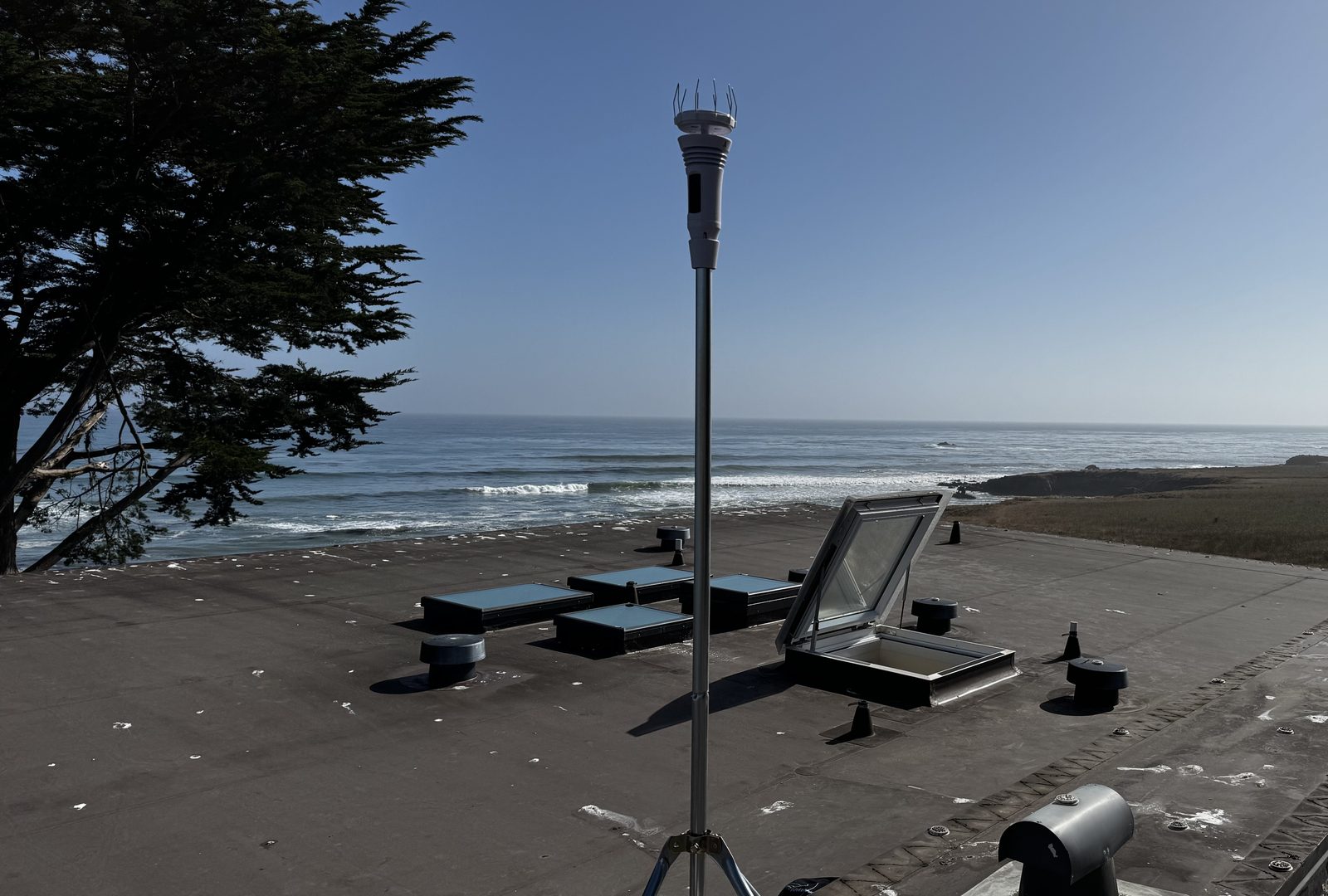 The WeatherFlow Tempest weather station mounted on a pole on the flat roof of Bluff House in Cayucos, with the Pacific Ocean and breaking surf in the background and a cypress tree to the left.